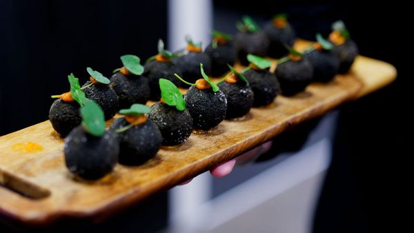 A wooden board with a row of black, spherical appetisers garnished with green leaves.