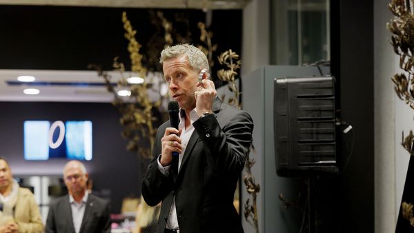 A man in a suit speaks into a microphone, holding a small object.