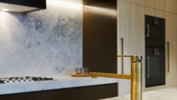 Bronze tap in foreground with a background of Gaggenau appliances installed in a bright luxury kitchen