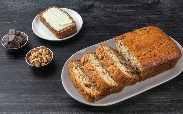 A sliced banana bread, along with two small containers of chocolate chips, walnuts and a buttered slice of banana bread.