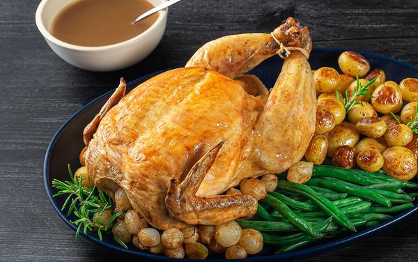 A succulent roast chicken is served on dark dish with green beans, potatoes and rosemary.