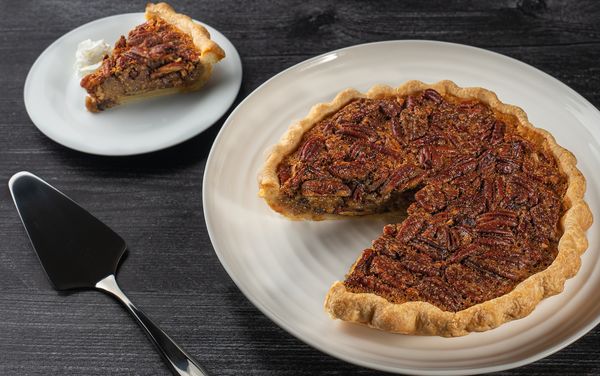 A pecan pie is shown with a slice missing, the slicing utensil and slice of pie is shown just out of shot.