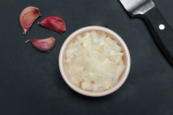 A stone worktop with three cloves of garlic and a white ramekin containing a chopped shallot