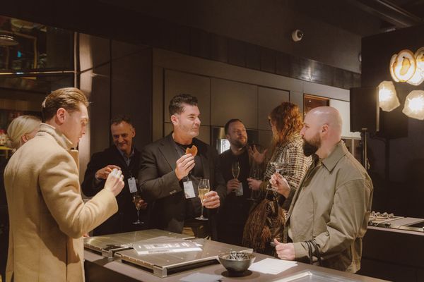Attendees holding drinks and food while engaging in conversations in an elegant kitchen setting with Gaggenau appliances visible.