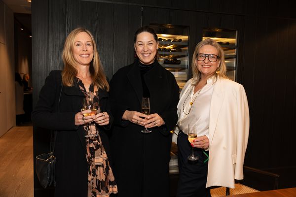 Three women smile together, holding glasses of wine during a sophisticated evening event.