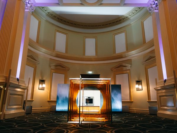 A modern Gaggenau kitchen display framed in gold within a grand, dramatically lit event hall with classical columns.